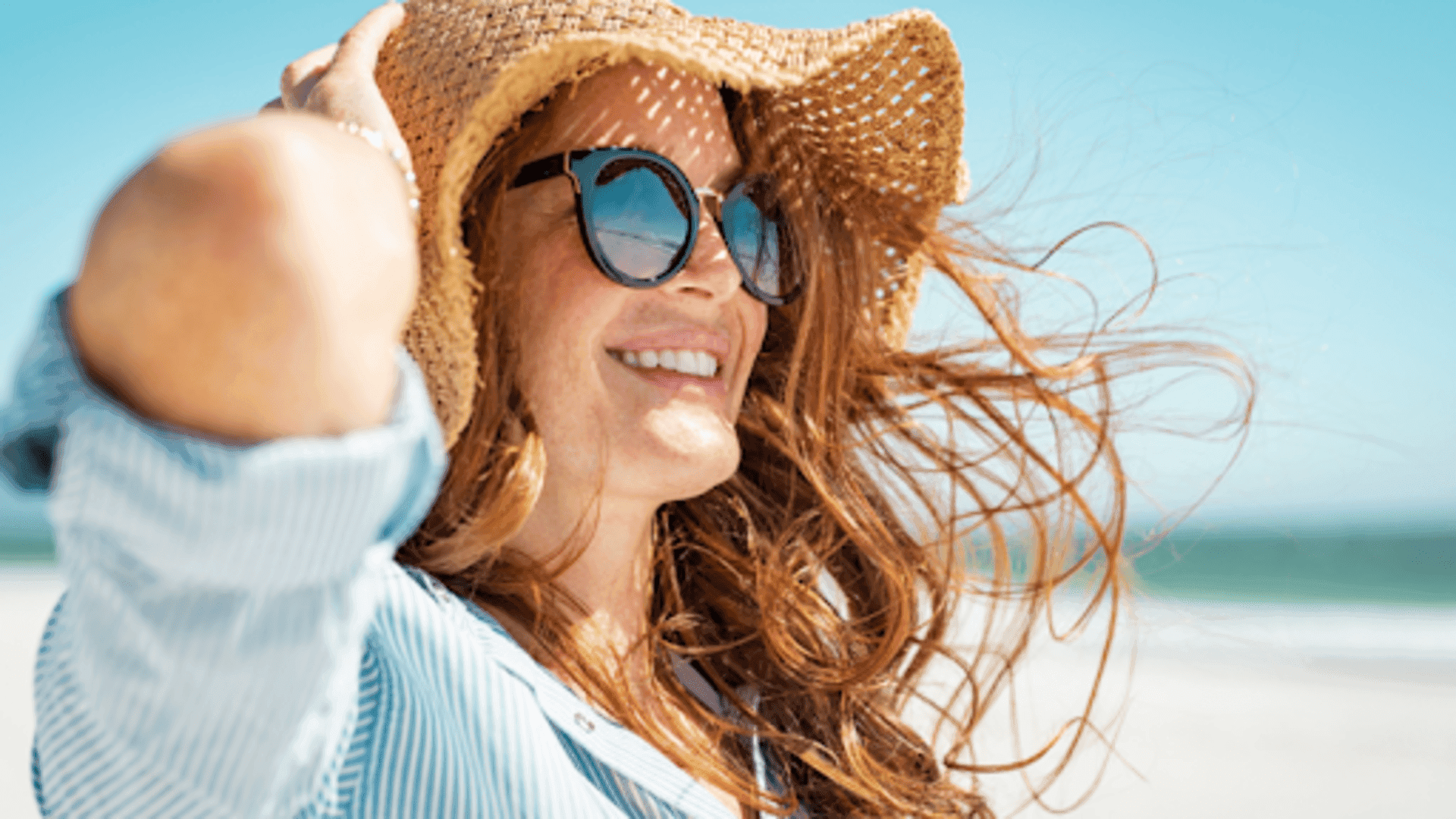 Mulher sorrindo sob o sol, usando óculos escuros e chapéu de palha, com cabelos ao vento em uma praia.
