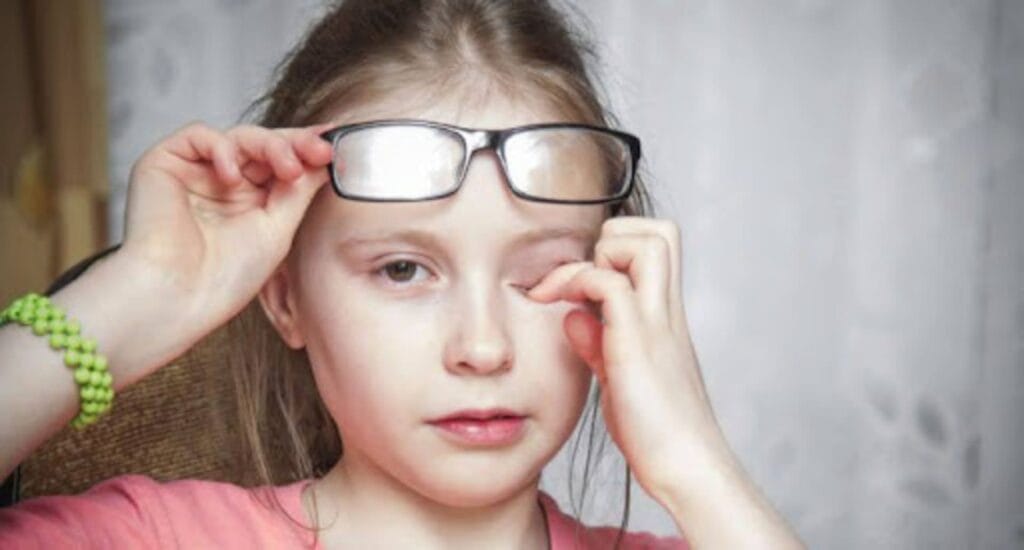 Menina ajustando os óculos e esfregando um dos olhos, aparentando desconforto visual.